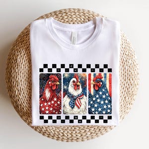 May include: White t-shirt featuring three rooster illustrations with patriotic themes. The roosters are depicted in red, white, and blue, with star and stripe patterns. The design is framed by a black and white checkered border.