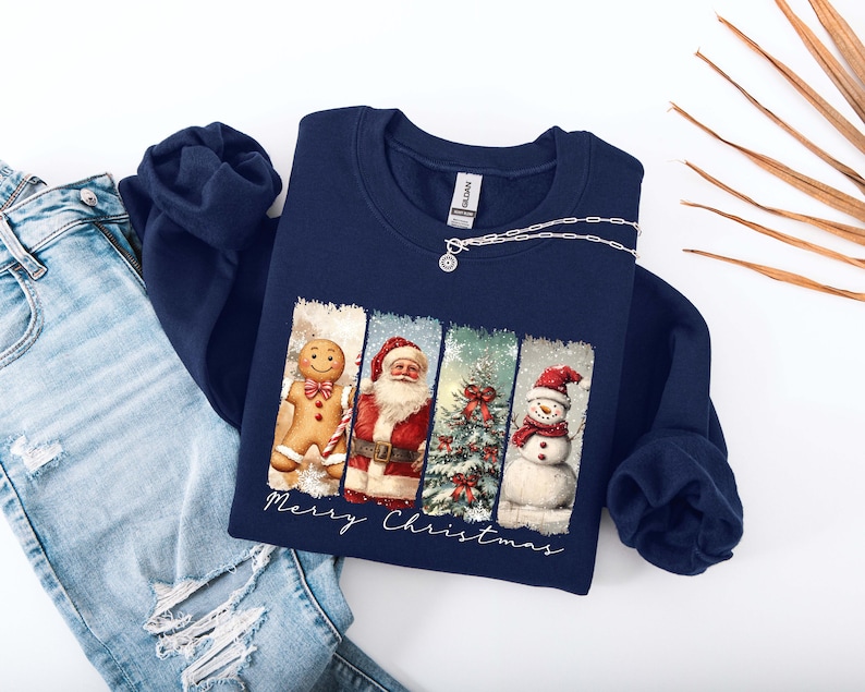 Puede incluir: Sudadera azul marino con un dise&ntilde;o navide&ntilde;o. El gr&aacute;fico presenta un hombre de jengibre, Pap&aacute; Noel, un &aacute;rbol de Navidad y un mu&ntilde;eco de nieve. Las palabras "Merry Christmas" est&aacute;n escritas debajo. Un collar plateado con un colgante est&aacute; en la sudadera.