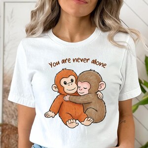 May include: White t-shirt featuring a cartoon illustration of two hugging monkeys. The monkeys are brown and orange, with the text "You are never alone" above them in brown lettering. The shirt is a casual style.