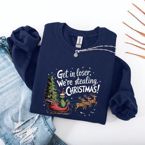 May include: Navy blue sweatshirt with the phrase "Get in loser, We're stealing CHRISTMAS!" printed in white. The graphic features a green character in a red sleigh with a Christmas tree and reindeer. A silver necklace is draped on the sweatshirt.