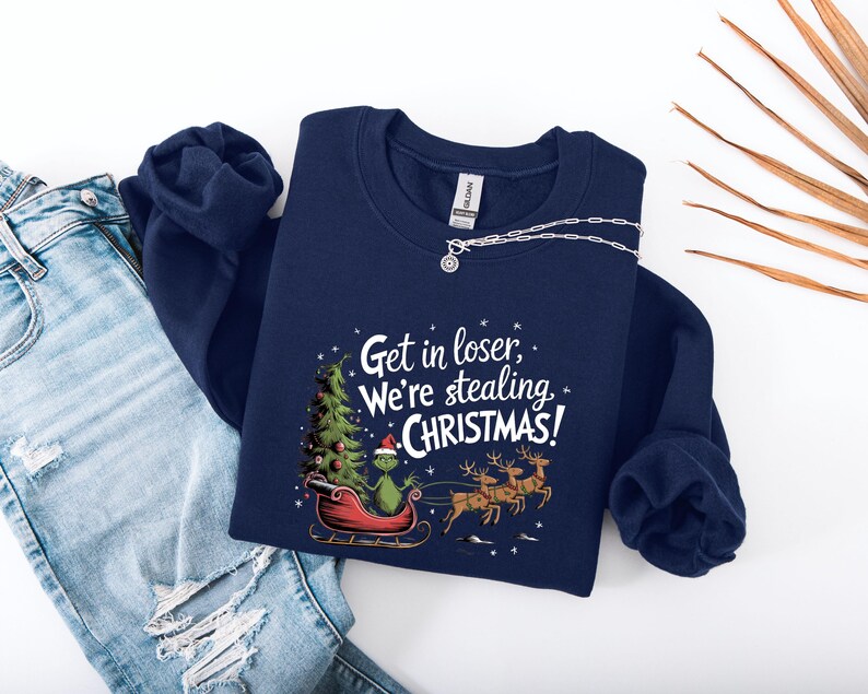 May include: Navy blue sweatshirt with the phrase "Get in loser, We're stealing CHRISTMAS!" printed in white. The graphic features a green character in a red sleigh with a Christmas tree and reindeer. A silver necklace is draped on the sweatshirt.