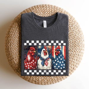 May include: Dark gray t-shirt featuring a patriotic design with three chickens. Each chicken is decorated with red, white, and blue colors and patterns, including stars and stripes. The shirt has a crew neck and short sleeves.