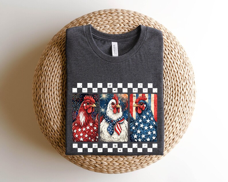 May include: Dark gray t-shirt featuring a patriotic design with three chickens. Each chicken is decorated with red, white, and blue colors and patterns, including stars and stripes. The shirt has a crew neck and short sleeves.