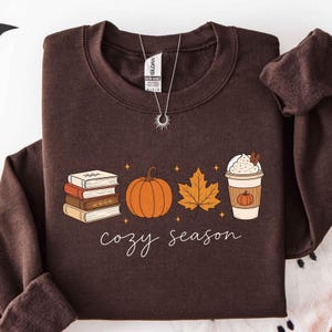 May include: A dark brown crewneck sweatshirt featuring a graphic print of a stack of four books, an orange pumpkin, a maple leaf, and a coffee cup with whipped cream. The text "cozy season" is printed in white cursive below the illustrations.