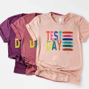 May include: Three t-shirts in shades of purple and pink. The front-most shirt is light pink with the words "TEST DAY" in colorful block letters. The shirt also has the text "You Got This!" and motivational phrases.
