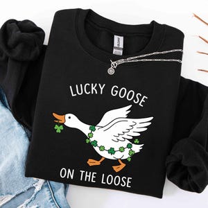 May include: Black sweatshirt with the text "LUCKY GOOSE ON THE LOOSE" and a cartoon goose wearing a shamrock garland and holding a shamrock in its beak. A silver necklace with a pendant is on the sweatshirt.