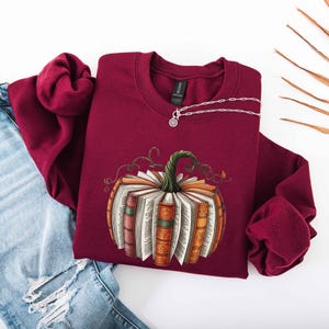May include: Burgundy crewneck sweatshirt with a pumpkin design made of books. The pumpkin has a green stem and is surrounded by vines. A silver necklace with a pendant is on the sweatshirt. Light blue jeans are on the left.