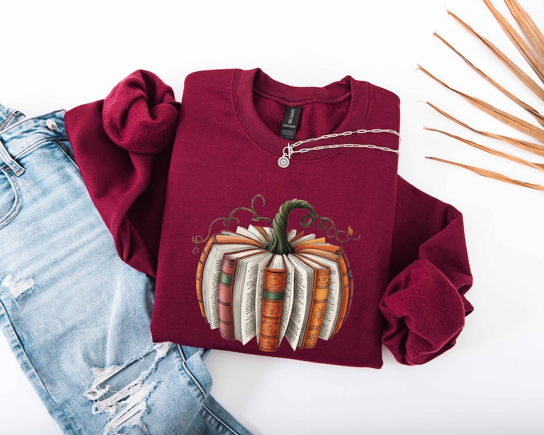 May include: Burgundy crewneck sweatshirt with a pumpkin design made of books. The pumpkin has a green stem and is surrounded by vines. A silver necklace with a pendant is on the sweatshirt. Light blue jeans are on the left.