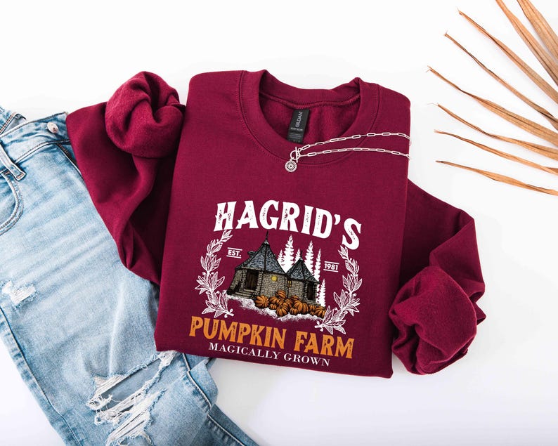 May include: A maroon sweatshirt with the text "Hagrid's Pumpkin Farm" in white, featuring a graphic of a house, trees, and pumpkins. A silver necklace is draped on the sweatshirt, which is next to a pair of blue jeans.