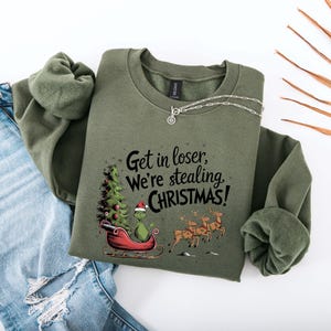 May include: An olive green sweatshirt with the text "Get in loser, we're stealing CHRISTMAS!". The graphic features a Grinch character in a sleigh with a Christmas tree and reindeer. A silver necklace and blue jeans are also visible.
