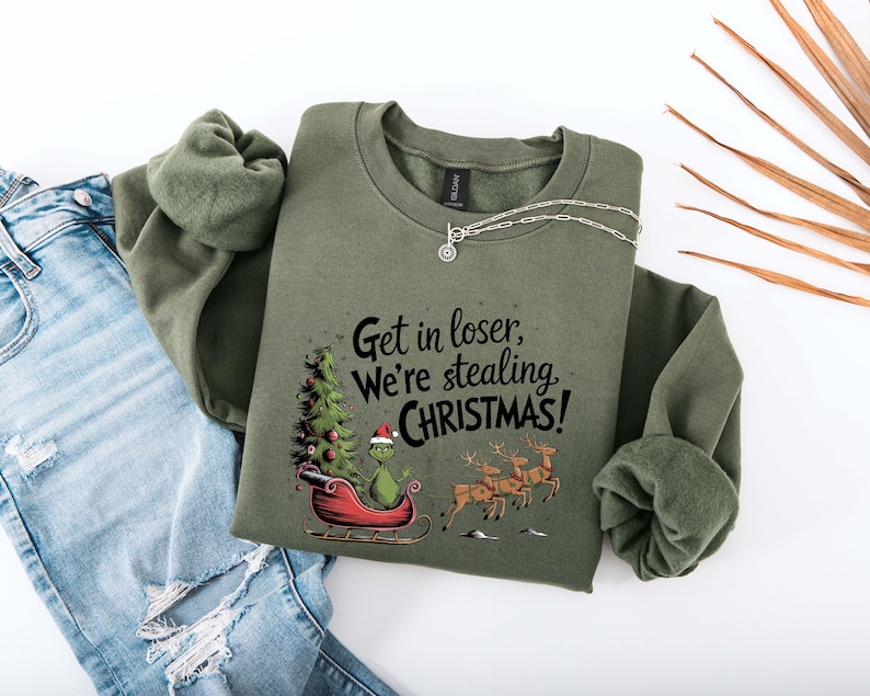 May include: An olive green sweatshirt with the text "Get in loser, we're stealing CHRISTMAS!". The graphic features a Grinch character in a sleigh with a Christmas tree and reindeer. A silver necklace and blue jeans are also visible.