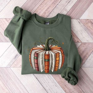 May include: An olive green sweatshirt featuring a pumpkin design made of open books. The pumpkin has a green stem and is composed of books with orange, red, and brown spines. The sweatshirt is folded on a light wood surface.