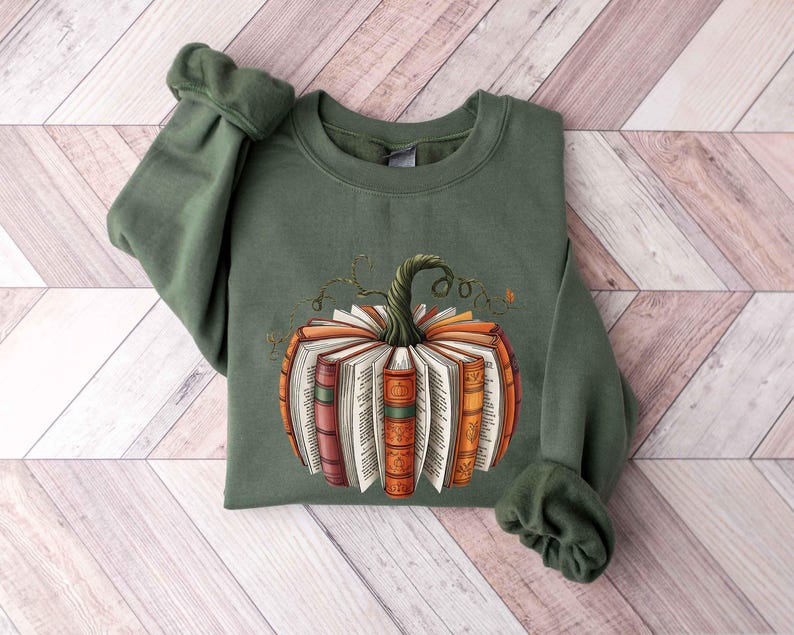 May include: An olive green sweatshirt featuring a pumpkin design made of open books. The pumpkin has a green stem and is composed of books with orange, red, and brown spines. The sweatshirt is folded on a light wood surface.
