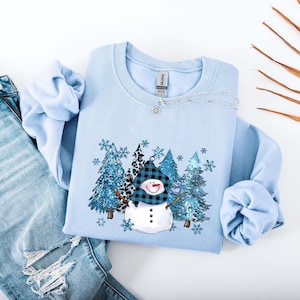 May include: A light blue sweatshirt featuring a snowman design with a black and blue plaid hat and a carrot nose. The graphic includes blue glittery trees and snowflakes. The sweatshirt is paired with light blue denim jeans.