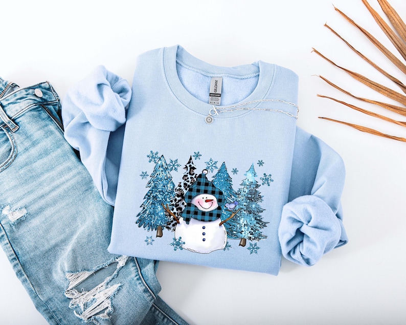 May include: A light blue sweatshirt featuring a snowman design with a black and blue plaid hat and a carrot nose. The graphic includes blue glittery trees and snowflakes. The sweatshirt is paired with light blue denim jeans.