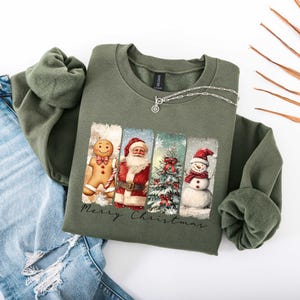 Puede incluir: Una sudadera verde oliva con un dise&ntilde;o navide&ntilde;o. Presenta un hombre de jengibre, Pap&aacute; Noel, un &aacute;rbol de Navidad y un mu&ntilde;eco de nieve. Las palabras "Merry Christmas" est&aacute;n impresas debajo. Un collar plateado est&aacute; sobre la sudadera.