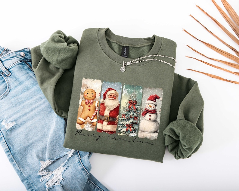 Puede incluir: Una sudadera verde oliva con un dise&ntilde;o navide&ntilde;o. Presenta un hombre de jengibre, Pap&aacute; Noel, un &aacute;rbol de Navidad y un mu&ntilde;eco de nieve. Las palabras "Merry Christmas" est&aacute;n impresas debajo. Un collar plateado est&aacute; sobre la sudadera.