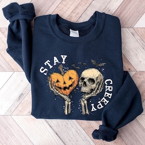 May include: Navy blue sweatshirt with a Halloween-themed graphic. The design features a carved pumpkin heart held by skeleton hands next to a skull, with the words "STAY CREEPY" in white. Two bats are also included in the design.