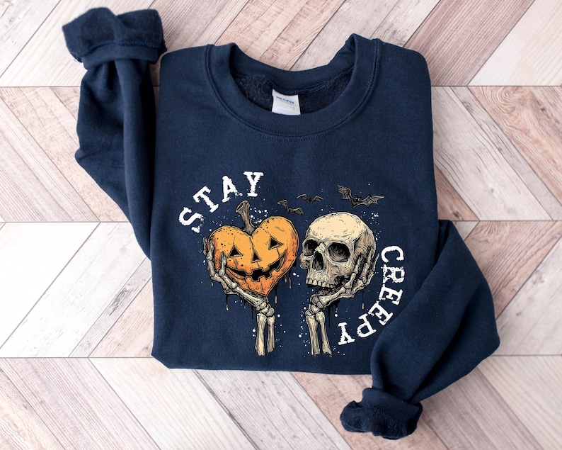 May include: Navy blue sweatshirt with a Halloween-themed graphic. The design features a carved pumpkin heart held by skeleton hands next to a skull, with the words "STAY CREEPY" in white. Two bats are also included in the design.