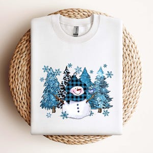May include: White sweatshirt featuring a snowman design with a blue plaid hat and scarf, and a smiling face. The design includes blue trees and snowflakes, creating a winter theme. The sweatshirt is displayed on a woven surface.
