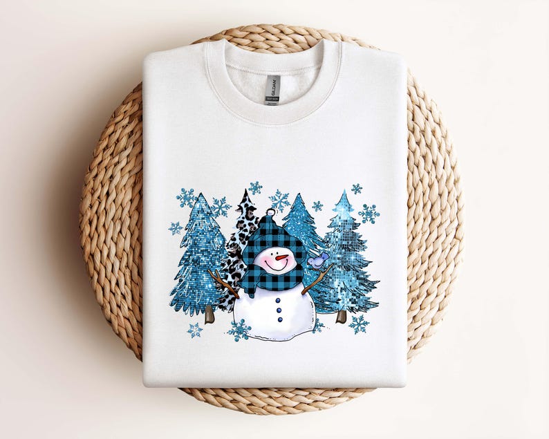 May include: White sweatshirt featuring a snowman design with a blue plaid hat and scarf, and a smiling face. The design includes blue trees and snowflakes, creating a winter theme. The sweatshirt is displayed on a woven surface.