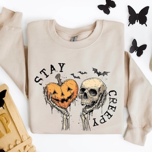 May include: Beige sweatshirt with a Halloween-themed graphic. The design features a jack-o'-lantern held by skeletal hands next to a skull, with the words "STAY CREEPY" and bat silhouettes. Decorative butterflies and a tombstone are also present.