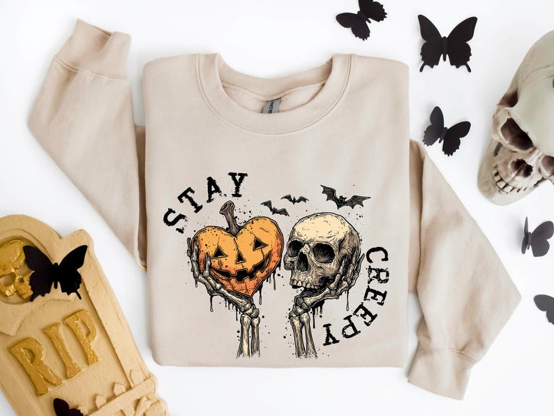 May include: Beige sweatshirt with a Halloween-themed graphic. The design features a jack-o'-lantern held by skeletal hands next to a skull, with the words "STAY CREEPY" and bat silhouettes. Decorative butterflies and a tombstone are also present.