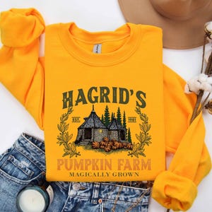 May include: Mustard yellow sweatshirt with a graphic that reads "Hagrid's Pumpkin Farm" with an illustration of a house, pumpkins, and trees. The text "Magically Grown" is below the farm name. The sweatshirt is folded on top of blue jeans.