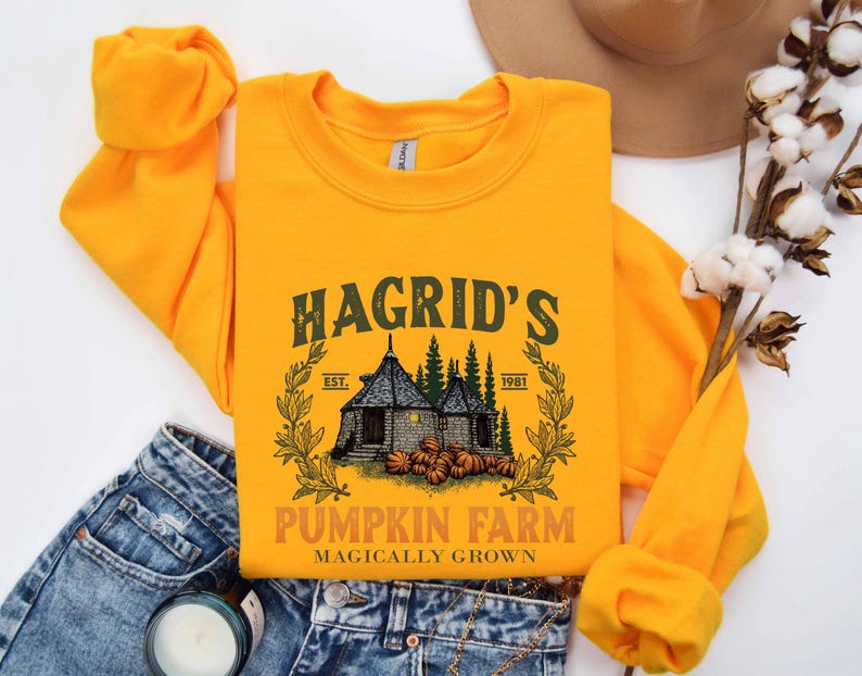 May include: Mustard yellow sweatshirt with a graphic that reads "Hagrid's Pumpkin Farm" with an illustration of a house, pumpkins, and trees. The text "Magically Grown" is below the farm name. The sweatshirt is folded on top of blue jeans.