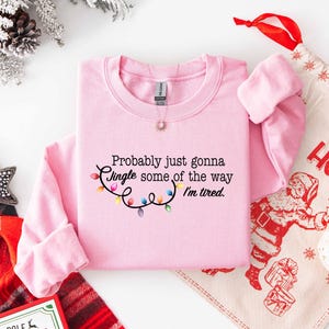 Puede incluir: Una sudadera rosa claro con el texto "Probably just gonna Jingle some of the way I'm tired." y una cadena de luces navide&ntilde;as de colores. Un tema festivo navide&ntilde;o con una bolsa de regalo navide&ntilde;a y decoraciones.
