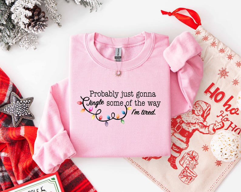 Puede incluir: Una sudadera rosa claro con el texto "Probably just gonna Jingle some of the way I'm tired." y una cadena de luces navide&ntilde;as de colores. Un tema festivo navide&ntilde;o con una bolsa de regalo navide&ntilde;a y decoraciones.