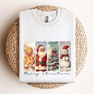 Puede incluir: Sudadera blanca con un dise&ntilde;o festivo de acuarela. Presenta un hombre de jengibre, Pap&aacute; Noel, un &aacute;rbol de Navidad y un mu&ntilde;eco de nieve, con "Merry Christmas" escrito debajo. La sudadera se muestra sobre una superficie circular tejida.