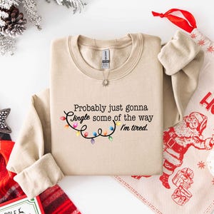 Puede incluir: Una sudadera beige con el texto "Probably just gonna Jingle some of the way I'm tired." El texto va acompa&ntilde;ado de una cadena de luces navide&ntilde;as de colores. Una bolsa de regalo y decoraciones navide&ntilde;as est&aacute;n en el fondo.