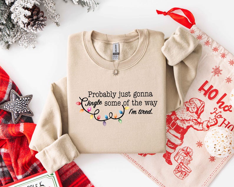 Puede incluir: Una sudadera beige con el texto "Probably just gonna Jingle some of the way I'm tired." El texto va acompa&ntilde;ado de una cadena de luces navide&ntilde;as de colores. Una bolsa de regalo y decoraciones navide&ntilde;as est&aacute;n en el fondo.