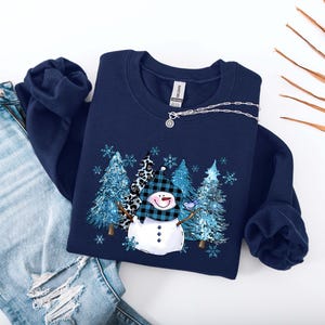 May include: Navy blue sweatshirt featuring a snowman design with a plaid hat and a carrot nose. The graphic includes blue trees and snowflakes. The sweatshirt is paired with distressed blue jeans and a silver necklace.
