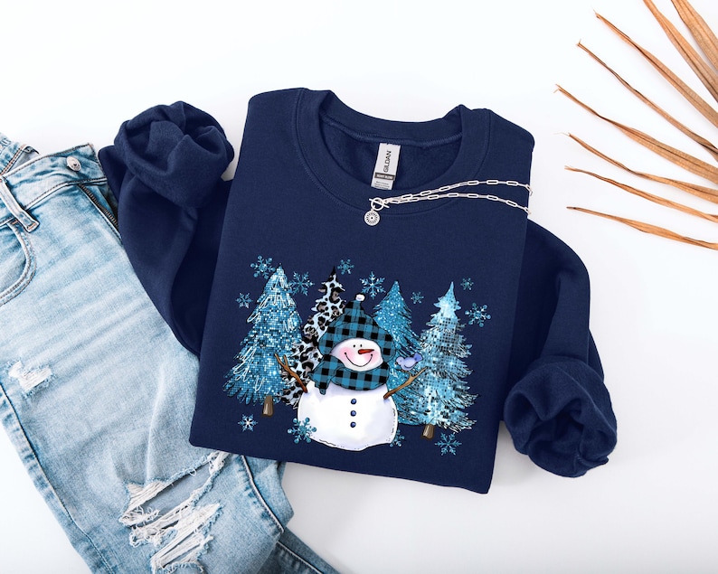 May include: Navy blue sweatshirt featuring a snowman design with a plaid hat and a carrot nose. The graphic includes blue trees and snowflakes. The sweatshirt is paired with distressed blue jeans and a silver necklace.