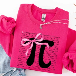 May include: A hot pink sweatshirt featuring a black Pi symbol with a pink bow. The Pi symbol is surrounded by a series of numbers. A silver necklace with a charm is draped across the sweatshirt.
