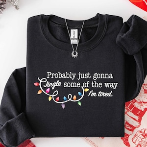 Puede incluir: Sudadera negra con cuello redondo con el texto "Probably just gonna jingle some of the way I'm tired." El texto es blanco con una cadena de luces navide&ntilde;as de colores. Se ve un collar plateado con un colgante de sol.