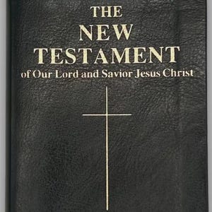 May include: A black book with the title "THE NEW TESTAMENT" in gold lettering, with the text "of Our Lord and Saviour Jesus Christ" below. A white cross is centered below the title. The book has a leather-like texture.