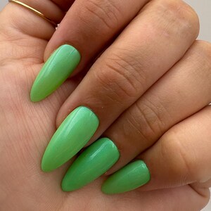 May include: A hand with long, almond-shaped, bright green artificial nails. The nails have a glossy finish and a vibrant, eye-catching color. A thin gold ring adorns the ring finger.