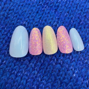 Könnte beinhalten: Fünf künstliche Fingernägel in einer Reihe, jeder mit einem anderen Design. Die Nägel sind hellblau, rosa mit Glitzer, gelb mit Glitzer, rosa mit Glitzer und hellblau. Die Nägel sind vor einem blauen Strickhintergrund angeordnet.