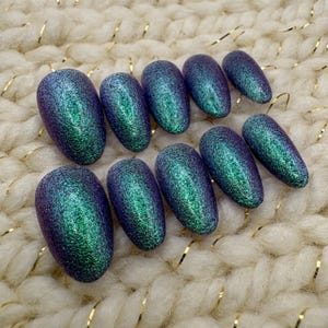 May include: A collection of ten oval-shaped, iridescent press-on nails. The nails shift from a deep purple to a vibrant green, producing a shimmering effect. The nails are displayed on a soft, off-white textured surface.