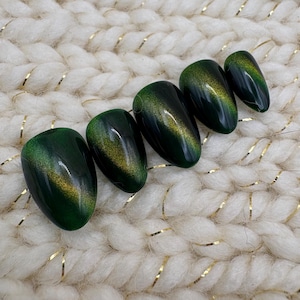 May include: Five oval-shaped, dark green press-on nails with a cat-eye effect. The nails have a glossy finish and a gold shimmer that runs horizontally across each nail. The nails are arranged on a white, textured surface.
