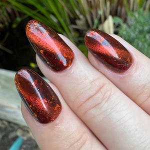 May include: Close-up of three fingernails painted with a dark red and orange metallic nail polish. The polish has a reflective, glittery finish with flecks of orange and gold. The nails are oval-shaped and have a glossy appearance.