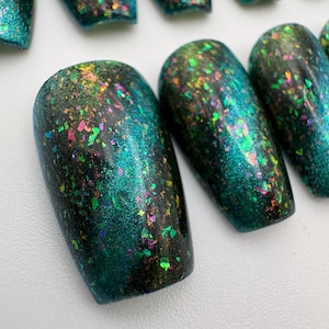 Op de afbeelding: Een set kunstnagels met een diepe blauwgroene en zwarte basiskleur en een afwerking van veelkleurige, iriserende vlokken. De vlokken glinsteren in tinten groen, roze en goud, wat zorgt voor een reflecterend en getextureerd uiterlijk op het oppervlak.