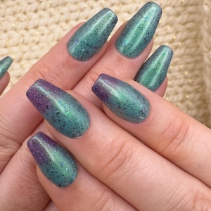 May include: Close-up of a hand displaying a set of long, coffin-shaped artificial nails. The nails feature a gradient design, transitioning from a deep purple at the base to a shimmering teal green at the tips. The nails have a glittery finish.
