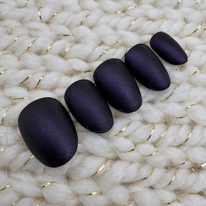 May include: Five matte, dark purple press-on nails arranged on a white, textured surface with gold accents. The nails have an oval shape and a smooth, non-glossy finish, perfect for a manicure.