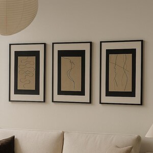 Puede incluir: Un salón moderno con un sofá color crema, una mesa de centro redonda de madera y tres impresiones artísticas abstractas enmarcadas. Una gran linterna de papel cuelga del techo. Los elementos decorativos incluyen un cojín marrón oscuro, un jarrón de cerámica y un cuenco pequeño.