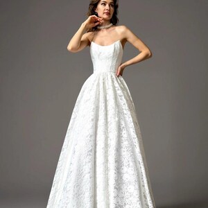 May include: A strapless white lace wedding dress with a full skirt. The dress features a fitted bodice and a delicate lace pattern. The model is wearing a pearl necklace. The dress is a classic bridal style.