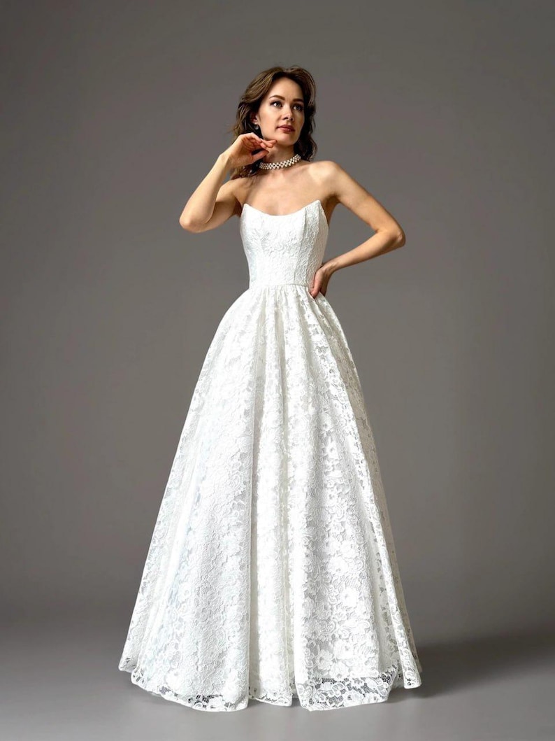 May include: A strapless white lace wedding dress with a full skirt. The dress features a fitted bodice and a delicate lace pattern. The model is wearing a pearl necklace. The dress is a classic bridal style.